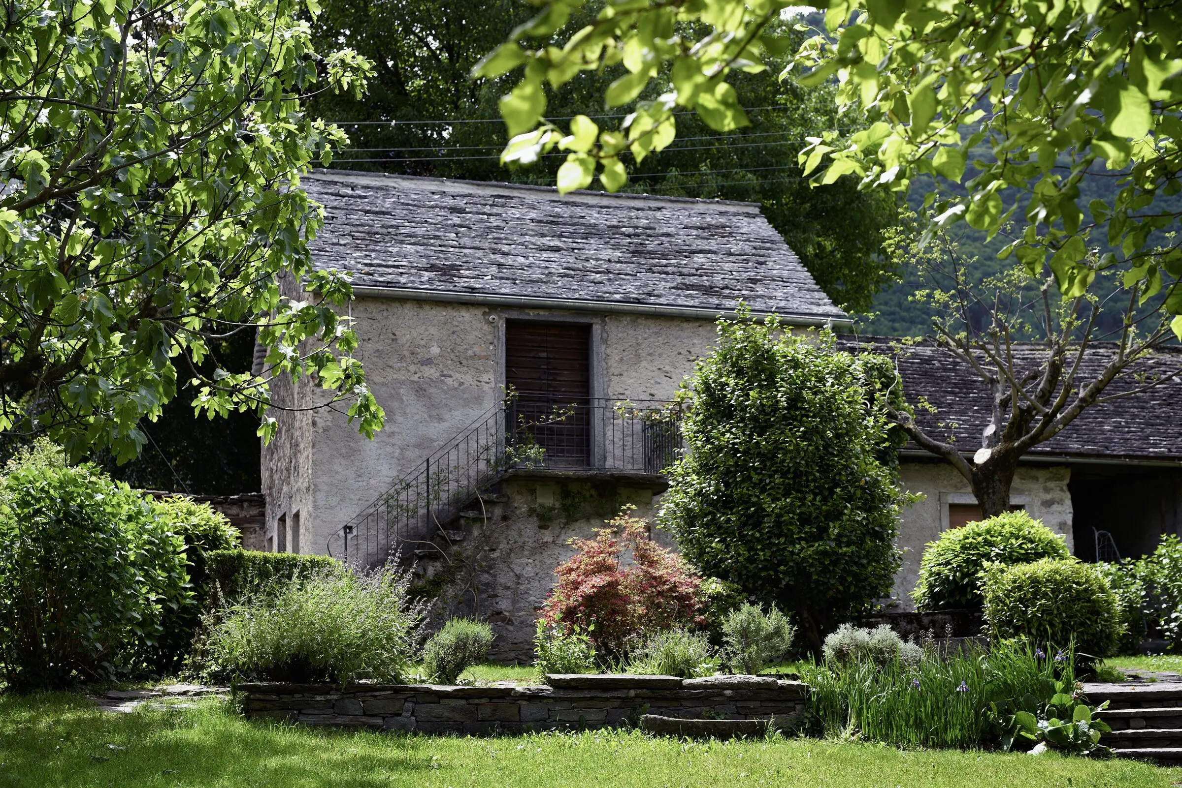 Steinernes Nebengebäude mit Balkon, umgeben von Bäumen und Beeten im Garten